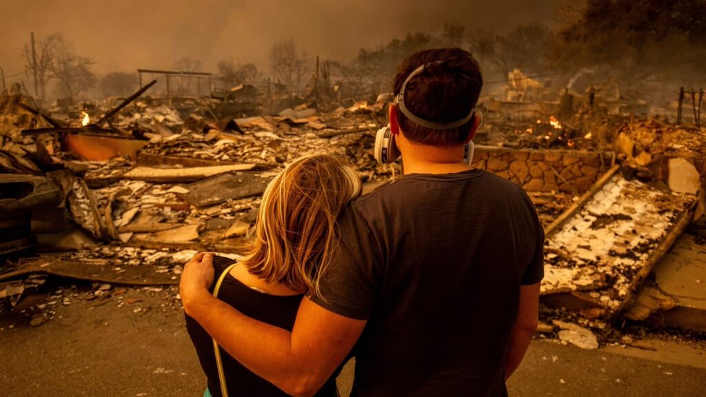 A man and woman standing in the foreground, looking out at rubble in the aftermath of a fire